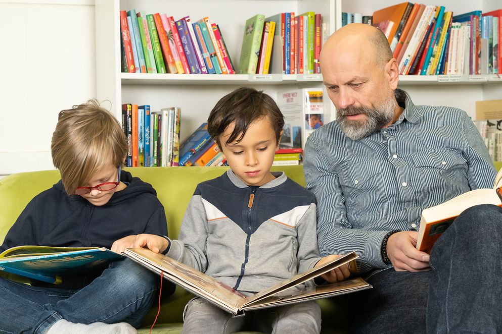 Zwei Kinder und ein Erwachsener sitzen lesend auf einer Couch